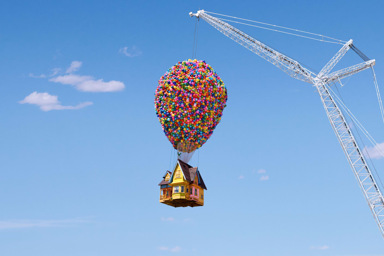A yellow house from the film Up hangs beneath a huge cluster of colorful balloons, suspended from a crane against a blue sky