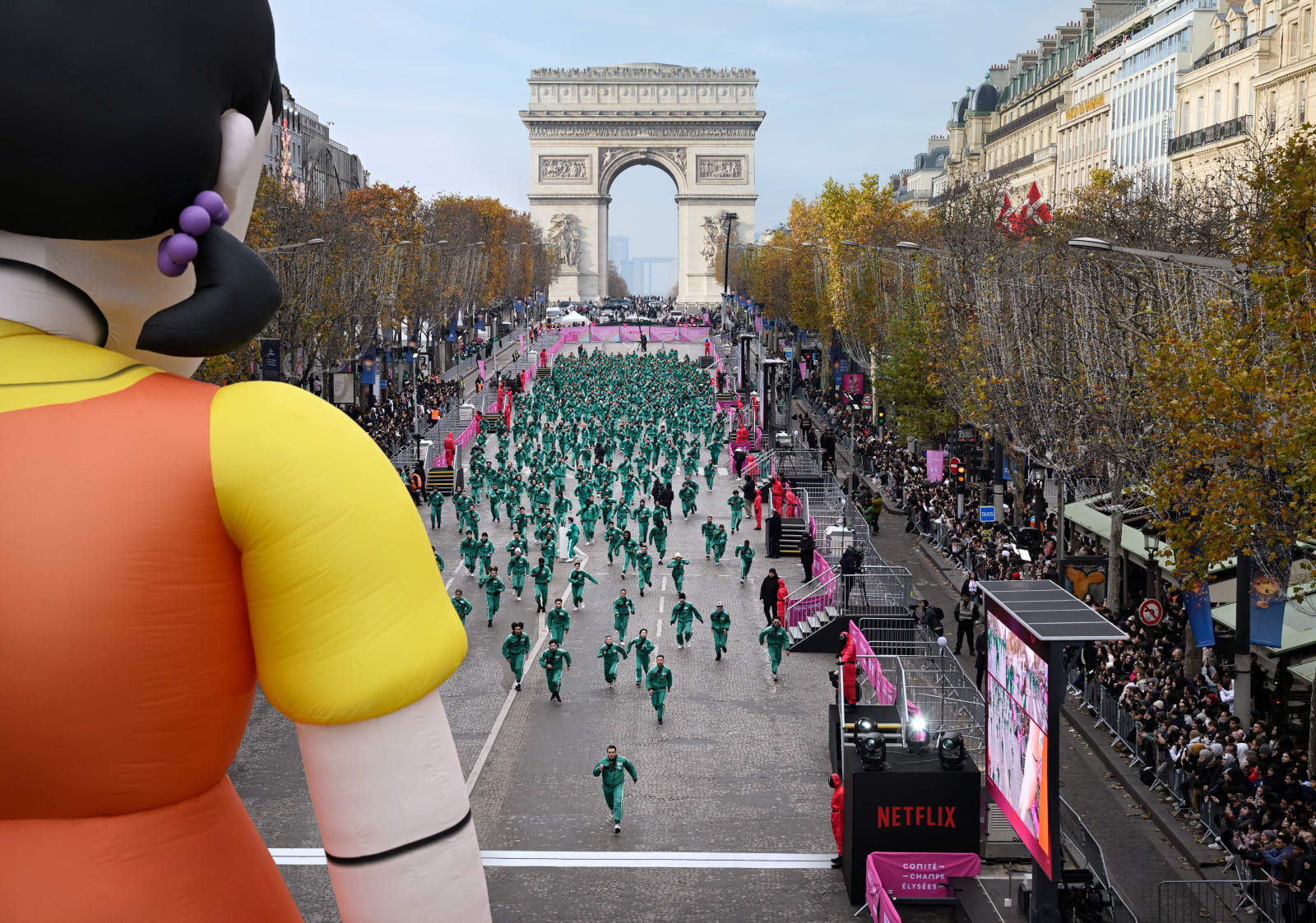 Players in green tracksuits run down the Champs-Élysées toward the Arc de Triomphe while a giant Young-hee doll watches