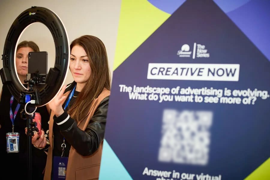 A guest poses at a ring-light tablet station beside a large branded sign that asks a question and shows a scan code for joining in
