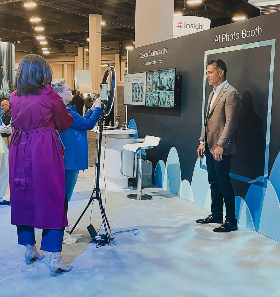 A staff member adjusts a phone on a ring light tripod while a guest poses in front of a branded trade show booth with a live display on the wall