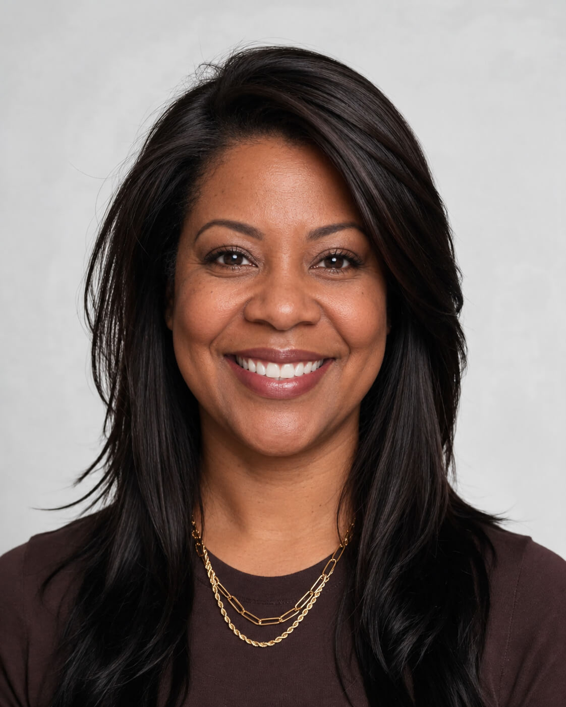 AI event headshot: smiling woman with long dark hair and layered gold necklaces against a light gray studio background